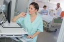 Bild på läkare eller sjuksköterska som sitter vid en dator. I bakgrunden syns en patient i en säng som pratar med en annan läkare.