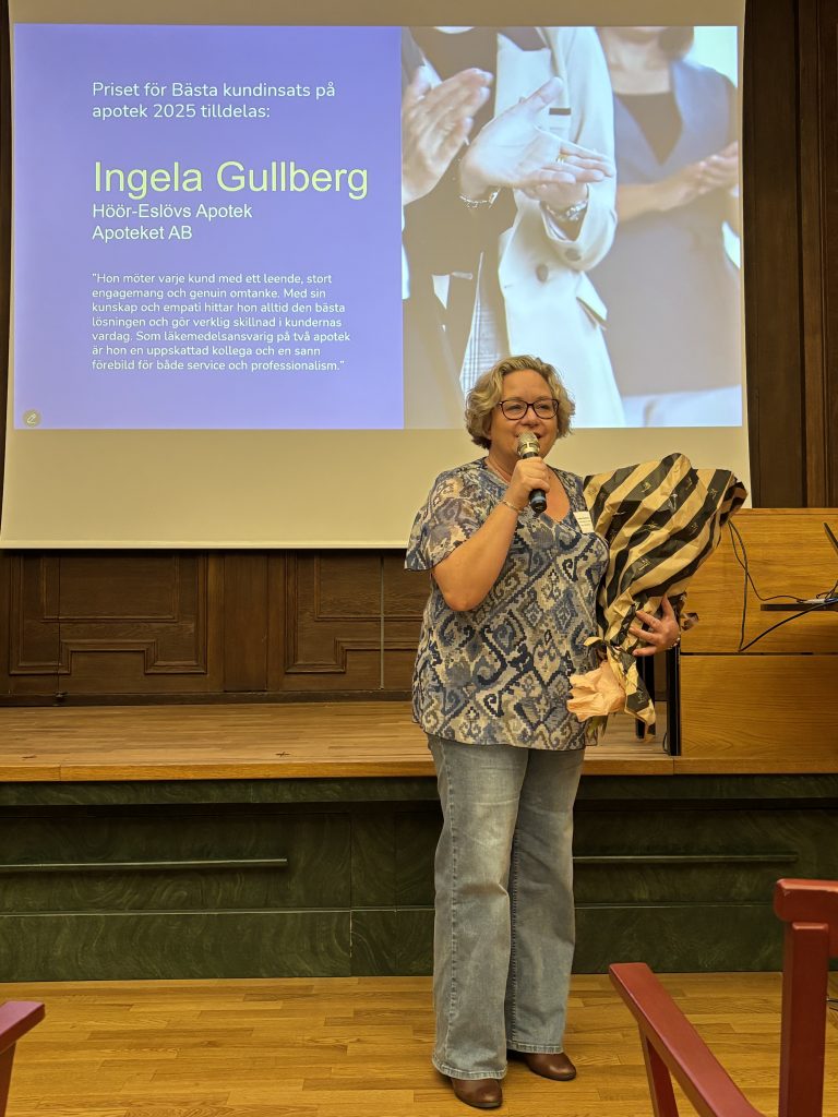 Ingela Gullberg står och talar i en mikrofon. Hon håller en bukett blommor i andra armen.