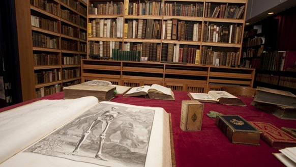 En öppen bok ligger på ett rött bord. I bakgrunden finns en bokhylla fylld med böcker.