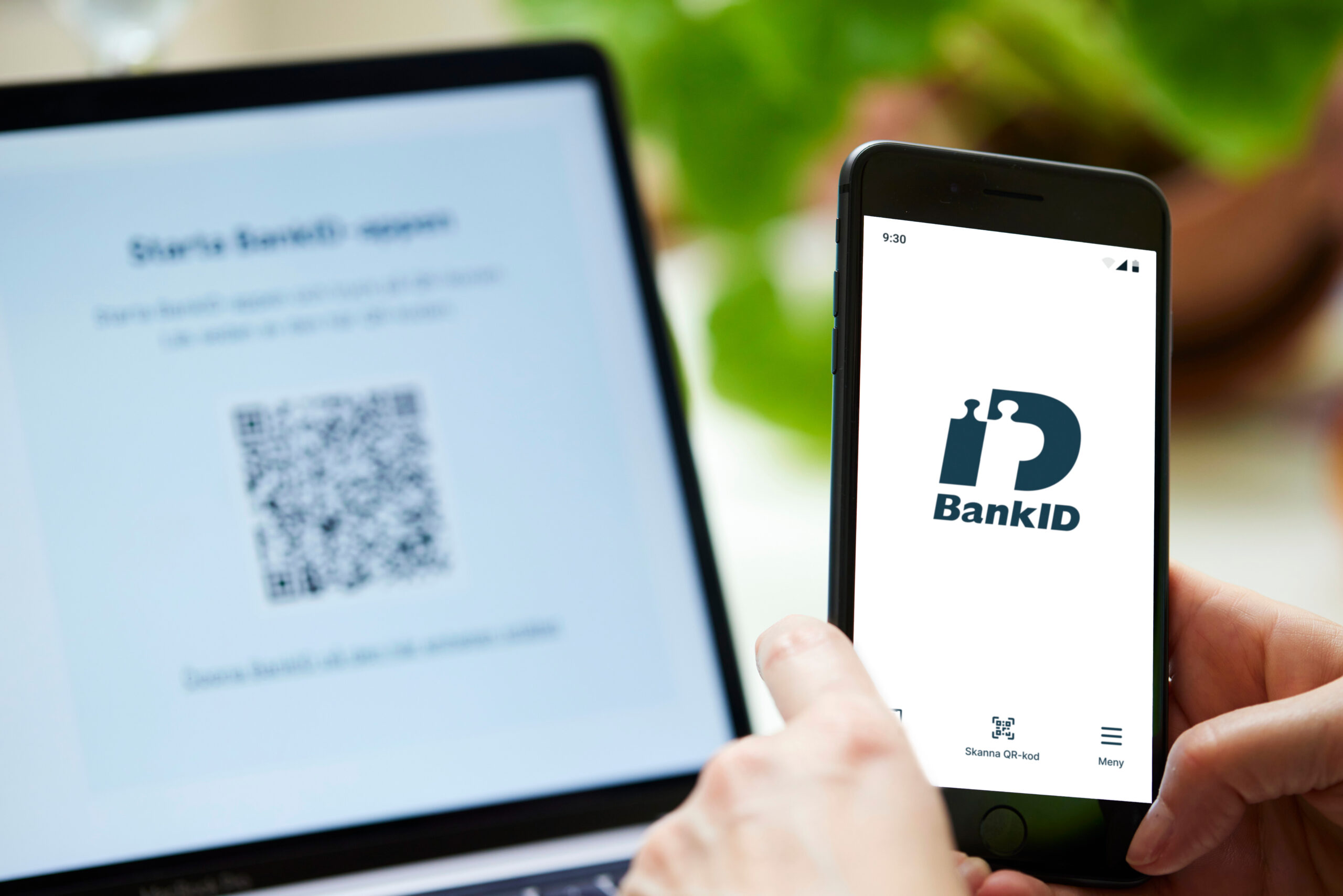 BankID-logotypen på en mobilskärm som hålls framför dator där QR-kod från BankID syns på skärmen.